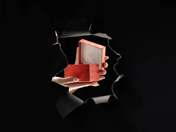 Holiday present. Black Friday. Night sale. Giveaway promotion. Female hands opening red gift box inside breakthrough paper hole ripped wall isolated on dark copy space background.