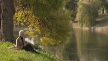 Sonbahar ilhamı. Romantik kadın. Keyifli vakit geçirme. Oldukça mutlu orta yaşlı bir bayan göl kıyısında oturuyor bir fincan kahve ve kitapla güzel doğanın tadını çıkarıyor..