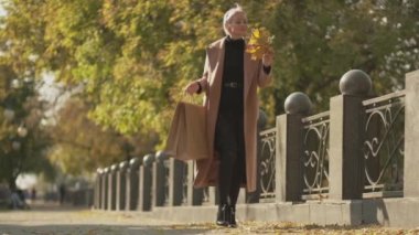 Mutlu sonbaharlar. Zarif kadın. Alışverişin tadını çıkarıyorum. Oldukça heyecanlı orta yaşlı bir kadın alışveriş torbalarıyla park yolunda yürüyor ve etrafa renkli yapraklar saçıyor..