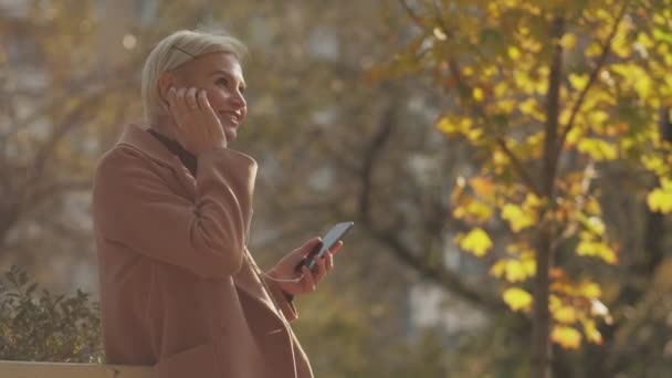 秋のインスピレーション 幸せな女性だ モバイル音楽 スマートフォンや有線のヘッドギアで公園を歩いている間 かなり興奮した中年の女性は好きな歌を聞いて — ストック動画