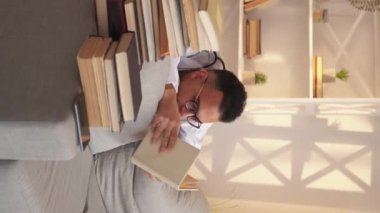 Dikey video. Doktor çalışması. Tıbbi araştırma. Bilgiye aşırı yüklenme. Yorgun stajyer çocuk, kitap yığınlarıyla kanepede ders çalışıyor..