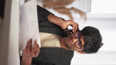 Dikey video. İş iletişimi. Ofis adamı. Rapor tartışması Kağıt döküman arayan gülümseyen adam akıllı telefon oturma odası lambasının iç kısmından bahsediyor..