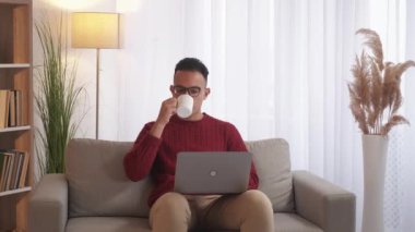 Çevrimiçi ev eğlencesi. Sanal sohbet. Dijital hafta sonu. Laptop kullanan rahat bir adam modern iç mekanda çay içiyor..