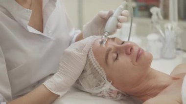 Yüz gerdirme prosedürü. Rahat kadın. Donanım kozmetolojisi. Profesyonel kozmetik uzmanı, orta yaşlı bayanın iç mekândaki alnına boşaltma masajı yapıyor..