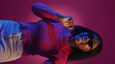 Dikey video. Parti eğlencesi. Kutlayan kadın. Neon ışıklı portre. Mor kırmızı arka planda dans eden güneş gözlüklü mutlu şık bayan yavaş çekim.