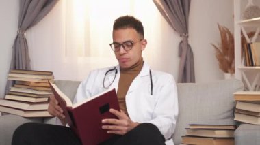 Sınav yorgunluğu. Tıbbi araştırma. Aşırı çalışma var. Yorgun erkek doktor stajyer profesyonel edebiyat okuyor kanepede kitap destesiyle modern ev içlerinde.