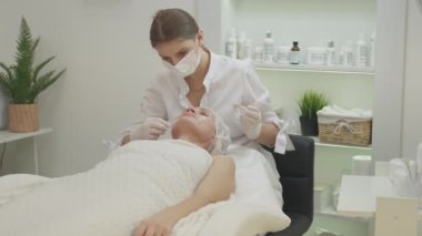 Güzellik prosedürü. Kadın yüz bakımı. Yenilenme kozmetiği. Kozmetik uzmanı, iç mekanda orta yaşlı bir kadına jel maske takıyor..