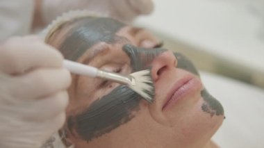 Yüz bakımı. Güzellik tedavisi. Salon prosedürü. Tanımlanamayan kozmetikçiler yüzlerine siyah kil macunu maskesi takıyor. Işıklı odada fırçası olan mutlu bir kadın..