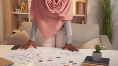 Mesafe çalışması. Konsantre kadın. Belgeleri kontrol et. Başörtüsü görünümlü ciddi bir bayan. Işık odasının iç tarafındaki masaya yayılan kağıt rapor..
