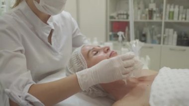 Güzellik prosedürü. Yüz bakımı. Yenilenme kozmetiği. Profesyonel kozmetik uzmanı, aydınlık odada orta yaşlı bir kadının cildini temizliyor..