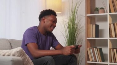 Gezici oyun. Sanal ev eğlencesi. Çevrimiçi eğlence. Neşeli, gevşemiş, gülümseyen adam modern oturma odasının iç tarafındaki koltukta akıllı telefondan eğleniyor..