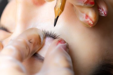 Dövmeci, modelin göz kapaklarını iki parmağıyla tutuyor ve silia arasında bir dövme yapıyor.