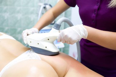 Kozmetoloji ofisindeki uzmanlar, beyaz iç çamaşırlarıyla kanepede yatan bir modele vakum masajı yapıyorlar.