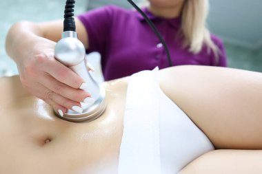 Beyaz iç çamaşırlı bir kız, karın bölgesindeki sorunlu bölgelerde ultrason makinesiyle ameliyat yapan bir ustanın önünde yatıyor.