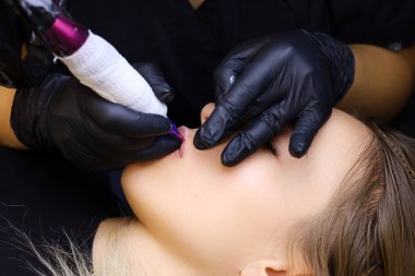 Dövmeci, dudaklarını tutarak modelin üzerine eğilir ve diğer eliyle dövme makinesiyle kalıcı makyaj yapar.