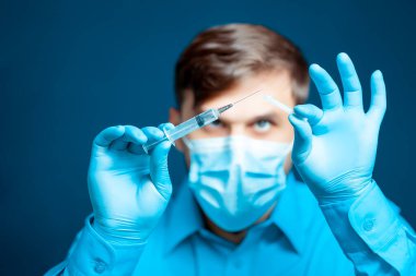 Tıbbi maskeli bir doktor ve mavi üniformalı bir eldiven şırınga iğnesinin kapağını çıkarıyor.