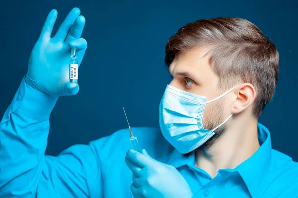 青い制服を着た医療用マスクと手袋の医者は注射器と彼の前でコロナウイルスに対するワクチンの入ったアンプルを持っています — ストック写真