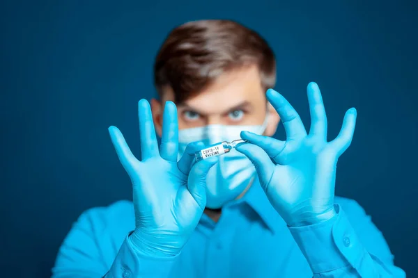 青い制服を着た医療マスクと手袋の医者は 彼の指で彼の前にコロナウイルスワクチンでアンプルを保持しています — ストック写真