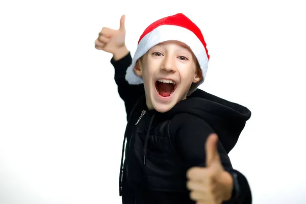 一个戴着红色圣诞帽 站在白色背景上的男孩竖起大拇指大笑起来 — 图库照片