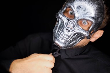 Siyah elbiseli bir adam kameranın önünde duruyor. İskelet maske takıyor ve kravatını düzeltiyor.