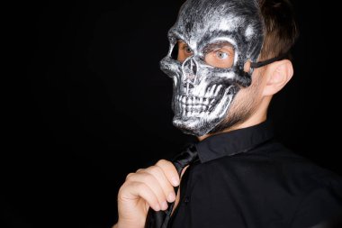 Siyah elbiseli bir adam kameranın önünde duruyor. İskelet maske takıyor ve kravatını düzeltiyor.