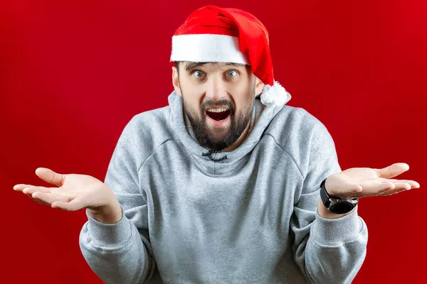 一个头戴红色圣诞老人帽的男人把胳膊伸向两边 迷惑不解地张开嘴 — 图库照片