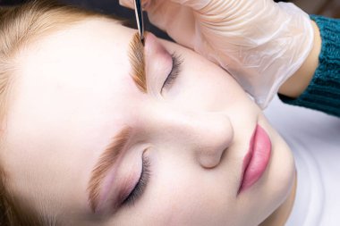 Ustanın elleri bir fırça tutuyor ve saç modelinin kaşlarını boyuyor laminasyon işleminden sonra saçlarını boyuyor.