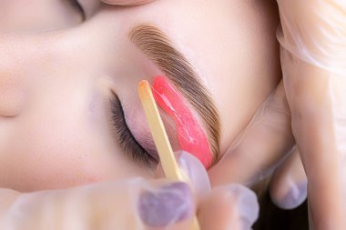 Usta beyaz eldivenlerin elleri modelin kaşlarını gerer istenmeyen kılları çıkarmak için renk ve kaplama prosedüründen sonra ağda uygularlar.