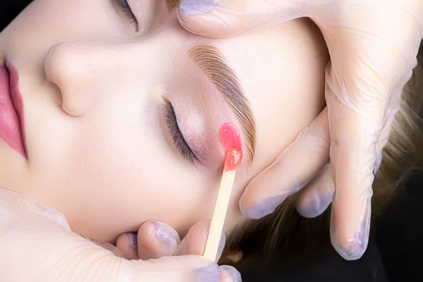 the master applies wax with a wooden stick to remove excess hairs from the lower contour of the eyebrows after the coloring and lamination procedure