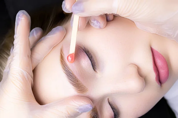 the master applies wax with a wooden stick to remove excess hairs from the lower contour of the eyebrows after the coloring and lamination procedure