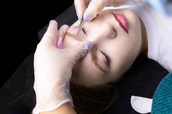 the hands of the master in white gloves stretch the eyebrow and pluck out excess hairs along the contour of the eyebrows