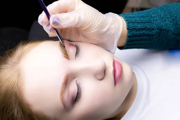 Ustanın elleri bir fırça tutuyor ve saç modelinin kaşlarını boyuyor laminasyon işleminden sonra saçlarını boyuyor.