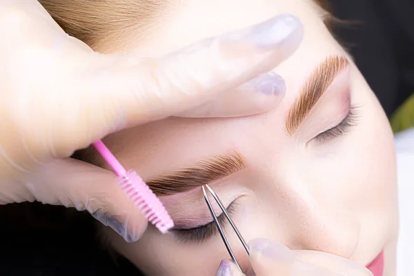 a close-up of the eyebrows of the model, the master holds in his hand a tweezer that removes excess hairs of already colored and laminated eyebrows