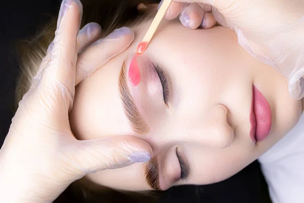 the master applies wax with a wooden stick to remove excess hairs from the lower contour of the eyebrows after the coloring and lamination procedure