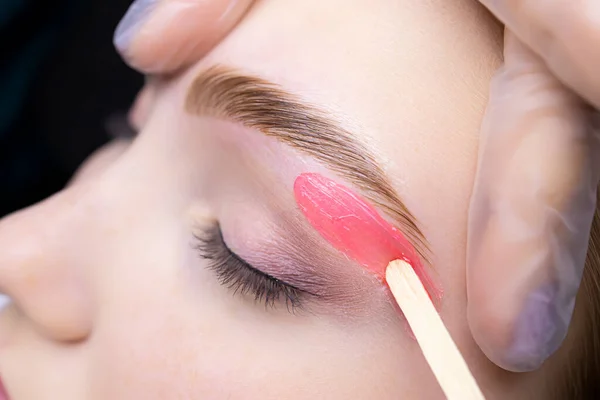 close-up of applying red wax to remove unwanted hairs from the lower contour of the model\'s eyebrows