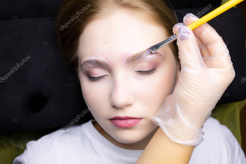 a young blonde model in the eyebrow lamination procedure, the master uses a brush to apply laminating compounds to the eyebrows