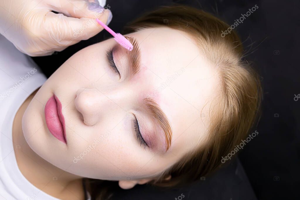 a close-up of the face of a blonde model who lies on the eyebrow lamination procedure, the master combs the eyebrows with a brush