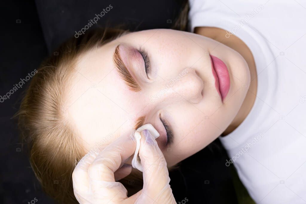 a close-up of the face of a blonde model who is lying on a black couch, the master wipes excess paint from her eyebrows with a cotton sponge