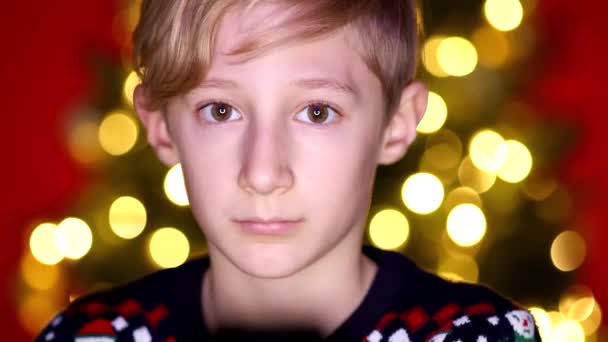 Een jongen in een kerst trui voor de camera achter hem is een kerst — Stockvideo