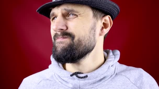 Een man met baard in een zwarte hoed op een rode achtergrond kijkt weg — Stockvideo