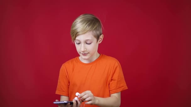 Pojke på en röd bakgrund håller en telefon i handen och sätter på hörlurar — Stockvideo