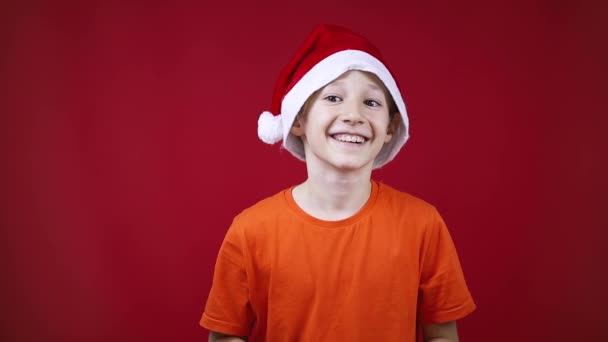 Niño vistiendo un sombrero de Navidad sonríe y se ríe en un fondo rojo a la cámara — Vídeos de Stock
