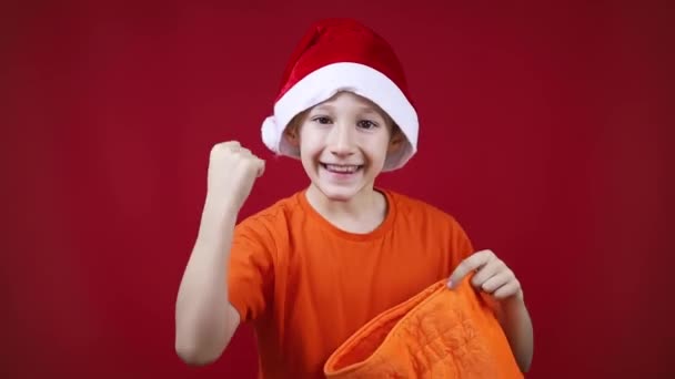 Een jongen in een rode kerstman kerstmuts houdt een geschenkzak in zijn handen — Stockvideo