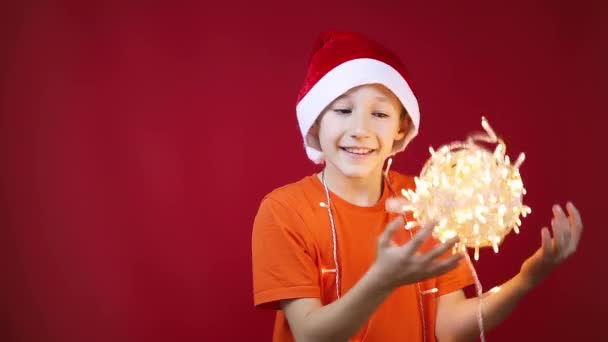 Un ragazzo con un cappello di Babbo Natale abbraccia ghirlande incandescenti — Video Stock