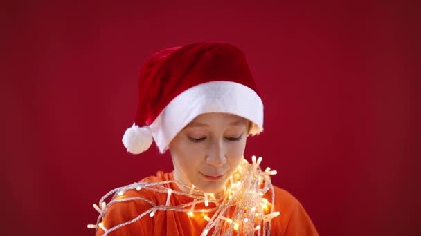 A boy in a Santas Christmas hat has a glowing garland around his neck — Stock Video