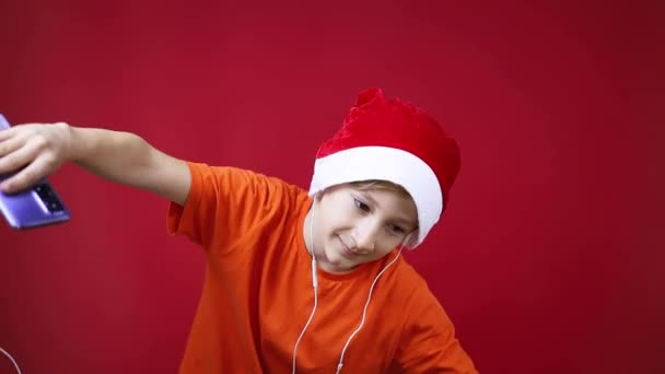 Chłopiec w świątecznym kapeluszu Mikołaja i żółtych koszulkach tańczy z telefonem — Wideo stockowe