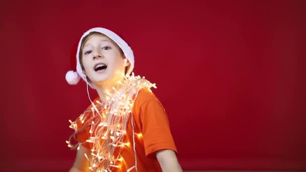 Een jongen op een rode achtergrond en een kerstmuts met een gloeiende kerst — Stockvideo