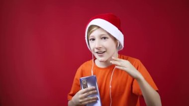 Kırmızı arka planda Noel Baba şapkalı bir çocuk kulaklarında kulaklıklarla dans ediyor.