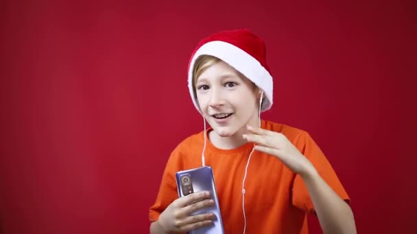 Un ragazzo con un cappello di Babbo Natale su uno sfondo rosso balla con le cuffie nelle orecchie — Video Stock