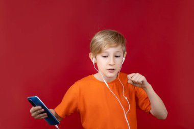 Kulağında kulaklığı olan neşeli bir çocuk bir telefon tutuyor ve kırmızı arka planda müzik dinliyor.